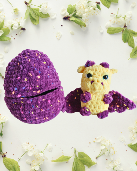 Crocheted dragon toy with a purple egg on a white background with green leaves and flowers.