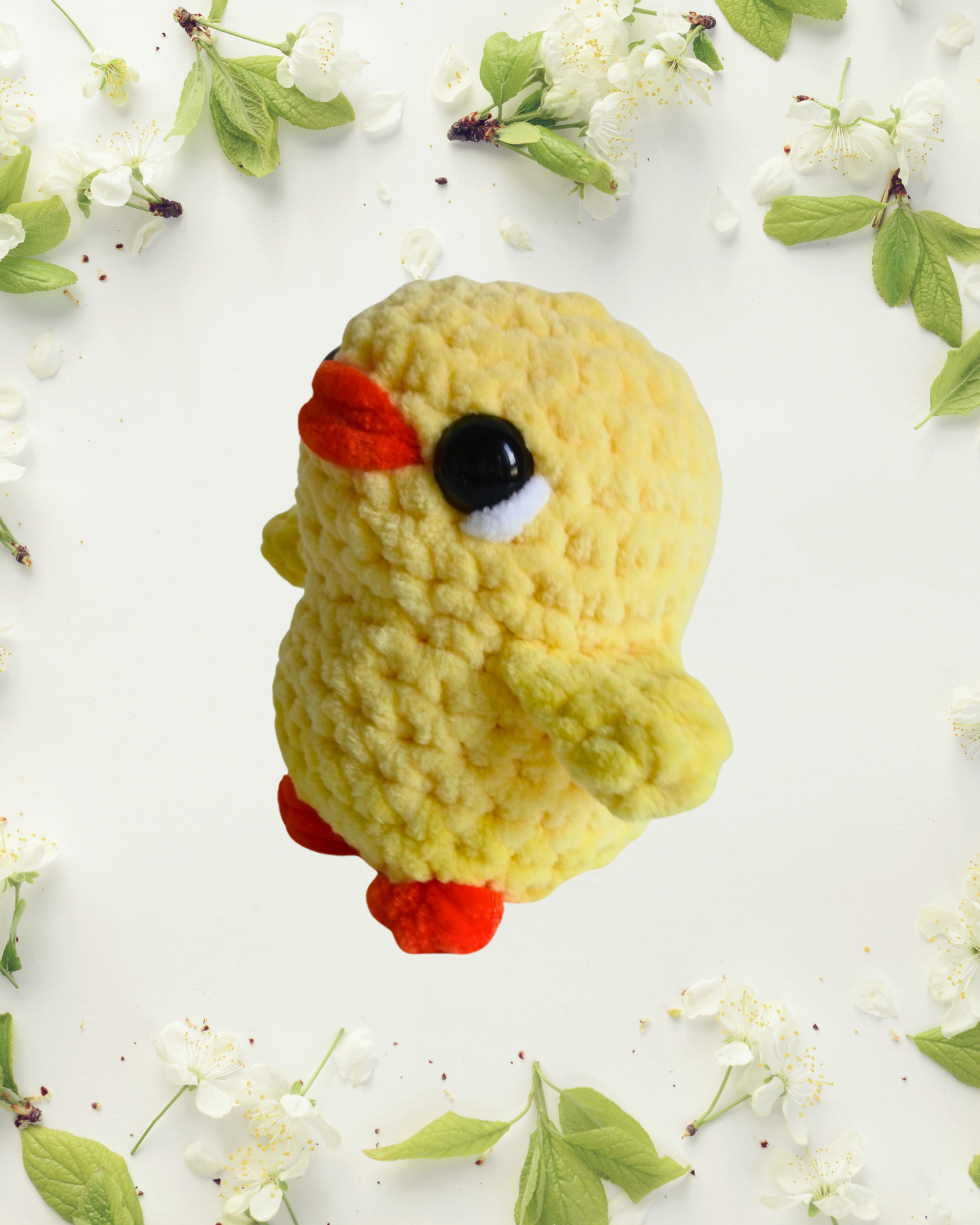 Crocheted yellow chick toy on a white background with green leaves and flowers.